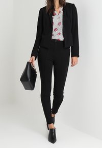 Blazer nero sopra una camicia bianca con motivi a pois e labbra, abbinato a pantaloni neri attillati e stivali neri con punta. Borsa nera.
