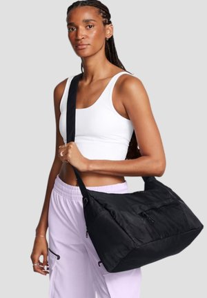 Donna con capelli intrecciati che indossa una canotta bianca e pantaloni viola chiaro, che porta una grande borsa nera a tracolla con tasca frontale in rete.