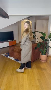 Trench coat beige lungo con cintura, pantaloni neri e scarpe con platform chiaro. In piedi in una stanza con pavimento in legno e piante.
