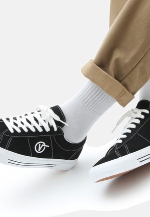 Schwarze Wildledersneaker mit weißen Schnürsenkeln und Nähten, mit einem Logo an der Seite. Tragen mit hellgrauen Socken und hochgekrempelten Khakihosen.