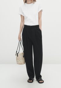 Vit T-shirt med uppvikta ärmar, svarta plisserade byxor, svarta sandaler och en vävd stråväska med svarta handtag; enfärgad bakgrund.