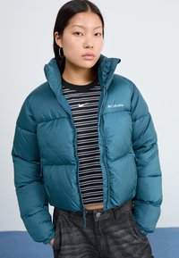 Teal pufferjakke med høy krage, glidelås foran og cropped design. Brukt over en svart-hvitt stripete skjorte.