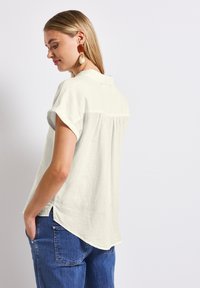 Lys beige kortærmet skjorte med krave, der har en subtil tekstur og en buet kant. Båret med blå jeans og accessoriseret med store øreringe.