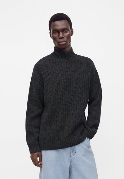 Homme à la peau foncée portant un pull à col roulé noir texturé et un jean bleu clair, debout devant un fond blanc uni.