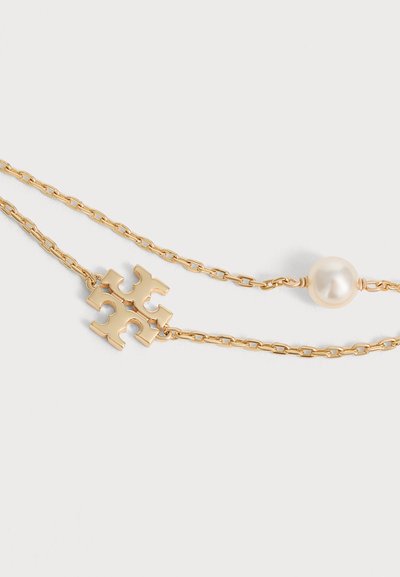 Collier chaîne en or avec un pendentif logo en or et une perle blanche ronde unique sur un fond clair uni.