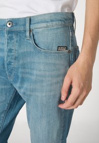 Ljusblå denimjeans med en slät yta, utrustade med knappstängning, framfickor och en G-Star-logopatch på midjan.