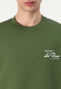 Olivgrüner Baumwoll-Sweatshirt mit geripptem Rundhalsausschnitt und einem weißen Aufdrucklogo auf der linken Brust. Schlichte, glatte Textur.