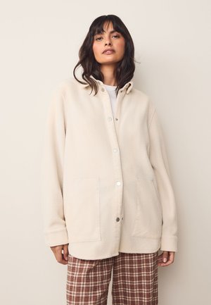 Giacca camicia in pile color crema con cappuccio, chiusura a bottone a pressione e due tasche frontali. Presenta polsini a coste e una texture morbida.