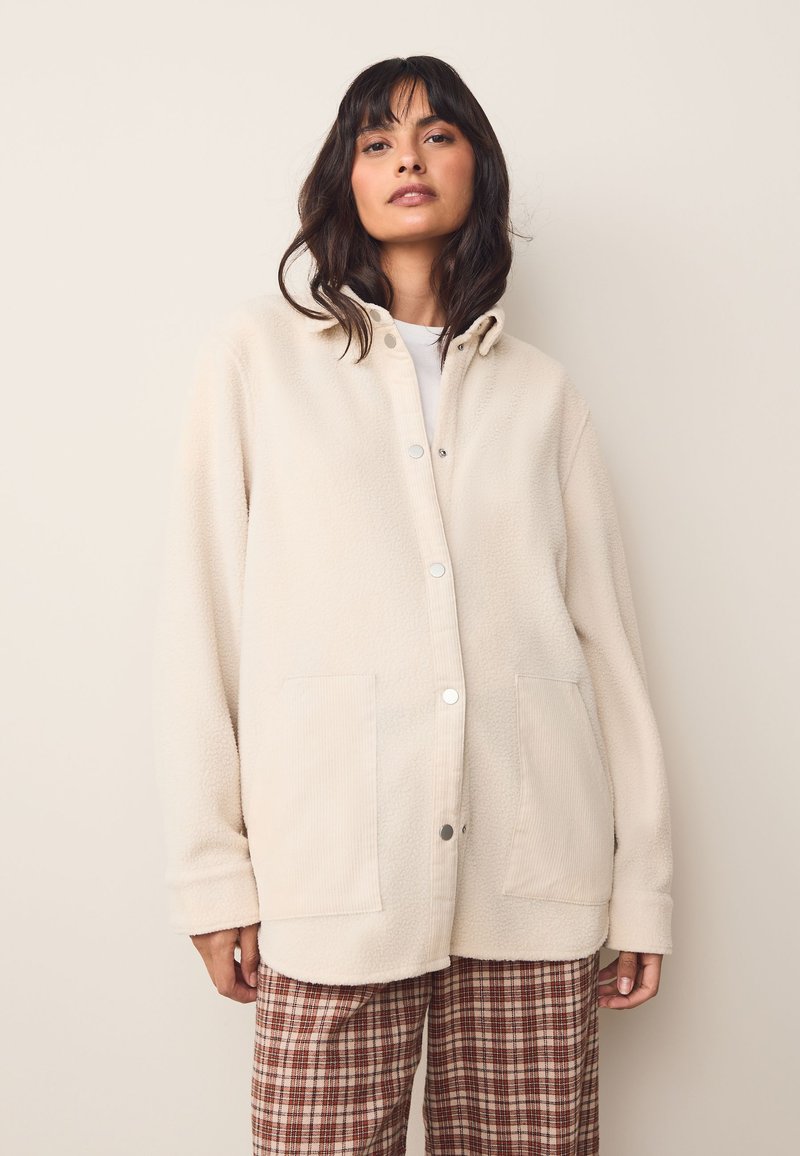 Giacca camicia in pile color crema con cappuccio, chiusura a bottone a pressione e due tasche frontali. Presenta polsini a coste e una texture morbida.