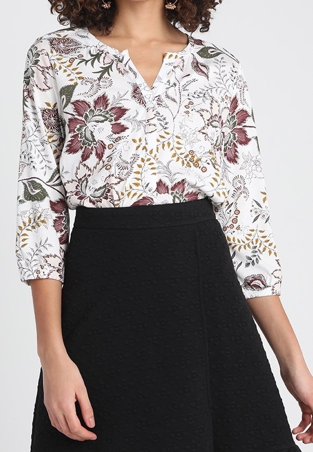 Florale Bluse in Weiß, mit roten, grünen und goldenen Mustern, V-Ausschnitt und Dreiviertelärmeln. Kombiniert mit einem strukturierten schwarzen Rock.
