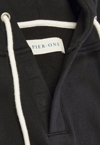 Felpa nera con cappuccio che presenta una patta cucita, un laccio bianco e un'etichetta con la scritta "PIER ONE" in testo blu. Tessuto interno testurizzato.