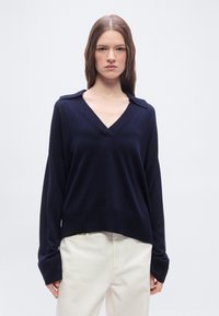 Νεαρή γυναίκα με μακριά καστανά μαλλιά, ντυμένη με μπλε σκούρο πουλόβερ με V λαιμόκοψη και εκρού παντελόνι, στέκεται μπροστά σε απλό ανοιχτό φόντο.
