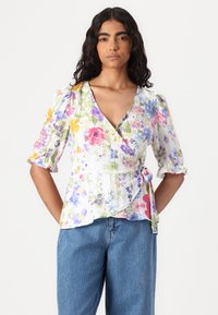 Blomsterinnpakningstopp i hvitt med lilla, rosa og gule mønstre. Har puffermer og en knytedetalj i livet. Kombinert med blå denimjeans.