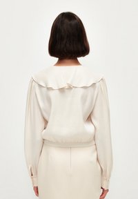 Blusa color crema con colletto a volant, maniche lunghe e vita elastica. Tessuto morbido e dettagli con bottoni ai polsini.