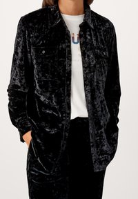 Giacca camicia in velluto nero con chiusura a bottoni, due tasche sul petto e vestibilità rilassata. Indossata sopra una T-shirt bianca con grafica.