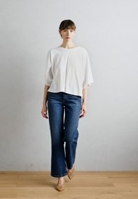 Blusa blanca de corte holgado con mangas anchas, combinada con jeans acampanados de color azul oscuro. La modelo lleva unos zapatos de tacón de yute en color beige. Fondo liso.