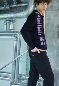 Cárdigan negro con patrón de punto geométrico morado, cierre de botón, usado sobre una camisa naranja. Combinado con pantalones negros que tienen un parche de logo.