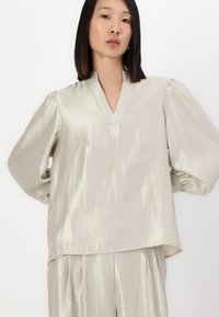Lys beige bluse med V-hals, puffede ærmer og en let skinnende tekstur. Har en lige skæring og er parret med matchende bukser.