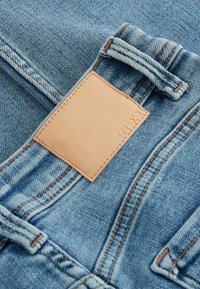 Světle modré džíny z denimu s hnědým koženým štítkem, viditelným šitím a smyčkovým detailem na pase. Hladká textura, tradiční design.