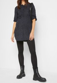 Schwarze Tunika mit Punkten, geraffter Taille, Dreiviertelärmeln, kombiniert mit schwarzen Leggings und klobigen schwarzen Ankle Boots.