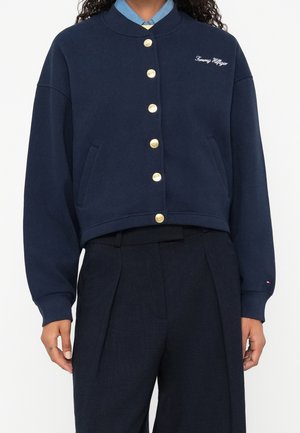 Person iført en marineblå knappet jakke med guldknapper og "Tommy Hilfiger" broderet på brystet over marineblå højttaljede bukser.