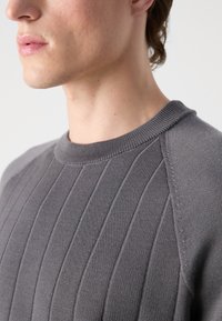Grau gerippter Pullover mit runder Ausschnitt und strukturierten Mustern entlang des Stoffes. Glatte Textur mit kontrastierenden Nähdetails.