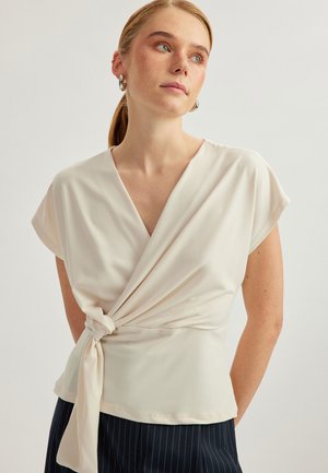 Top beige morbido con design avvolto, maniche corte e dettaglio con fiocco laterale. Realizzato in un tessuto leggero con una texture liscia.