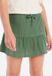 Falda mini verde con textura que presenta un diseño en capas, un patrón de ojales y una cinturilla ajustable con lazo. Adecuada para el clima cálido.