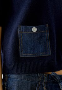 Navy gebreide trui met een denim zak voorzien van een strass drukknop. De zak heeft contrasterende gele stiksels. Ribgebreide zoomafwerking.