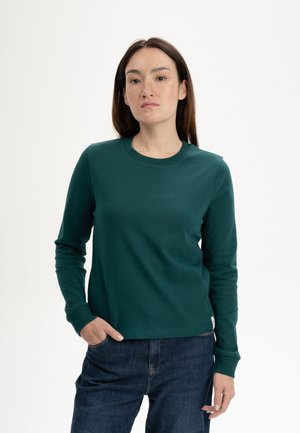 Frau mit glattem braunem Haar, die ein dunkelgrünes Langarmshirt und blaue Jeans trägt, mit einer Hand in der Tasche stehend vor weißem Hintergrund.