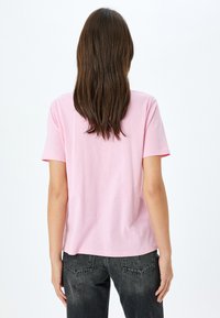 Camiseta corta de manga corta en color rosa claro, hecha de algodón, con un corte holgado y un cuello redondo, combinada con jeans oscuros, mostrada desde la parte de atrás.