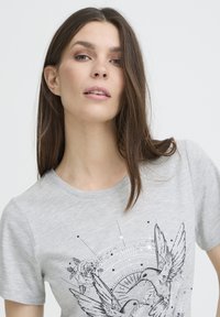 T-shirt gris à manches courtes avec un motif graphique d'un oiseau noir et d'une rose, des motifs circulaires et des accents en pointillés sur un tissu doux et texturé.