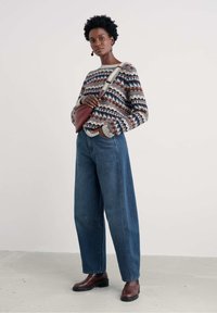 Moteris vilkinti raštuotą megztinį, plačius mėlynus džinsus, rudus aulinukus ir per petį nešamą rankinę, stovinti prieš paprastą foną.