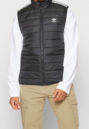 Mann trägt schwarze Adidas Steppweste über weißem Langarmshirt und beigen Cargohosen, steht vor einfachem Hintergrund.