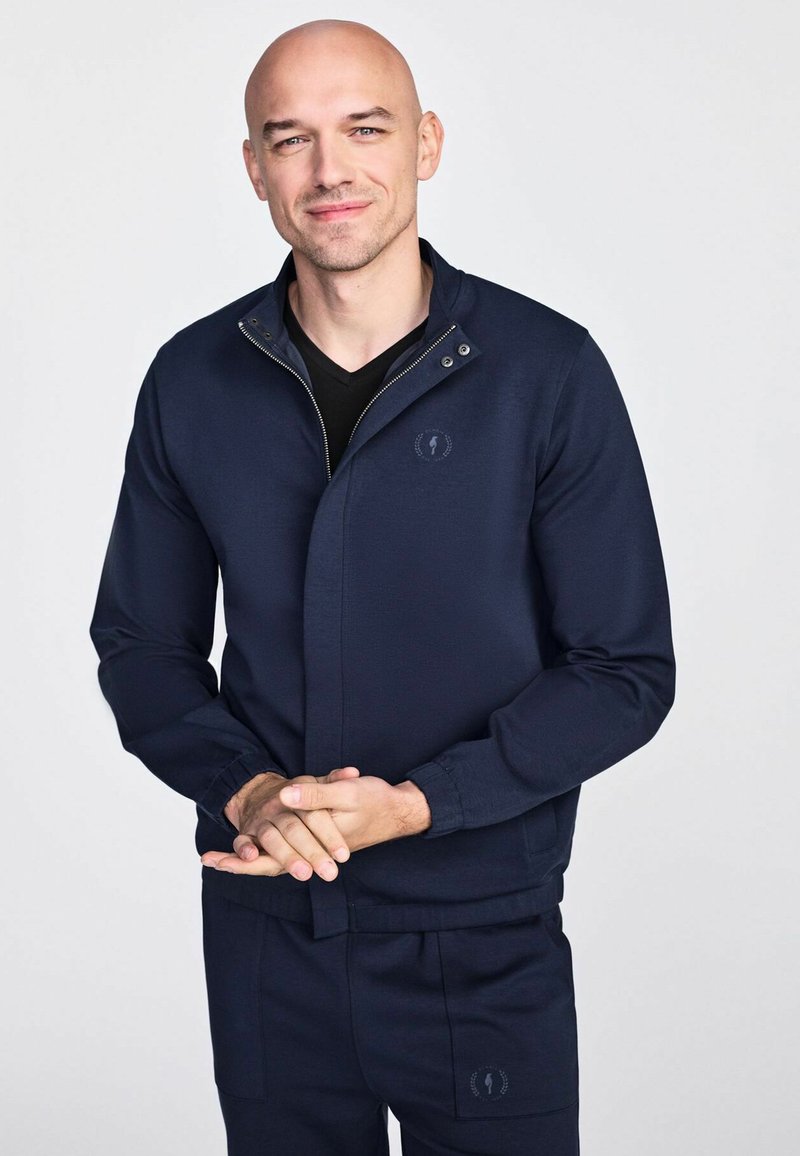 Granatowa kurtka zapinana na zamek z wysokim kołnierzem oraz detalem logo, charakteryzująca się gładką teksturą i dopasowanymi mankietami, w komplecie z pasującymi spodniami.