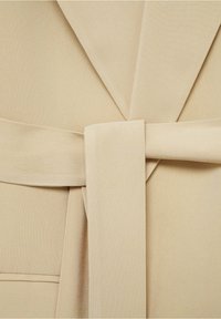 Beżowa marynarka z paskiem, wykonana z teksturowanej tkaniny, z wyraźnymi klapami i zachodzącymi na siebie panelami z przodu. Pasek jest związany w talii.