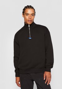 Black zip-up sweatshirt in soft fabric with high collar, featuring a blue logo tag and ribbed cuffs. Simple, minimalist design.