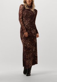 Frau in einem langärmeligen, bodenlangen Leopardenprint-Kleid, kombiniert mit schwarzen Stiefeletten, steht vor einem schlichten hellen Hintergrund.