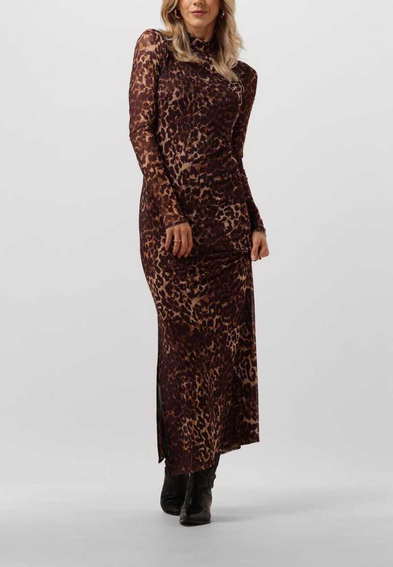 Frau in einem langärmeligen, bodenlangen Leopardenprint-Kleid, kombiniert mit schwarzen Stiefeletten, steht vor einem schlichten hellen Hintergrund.