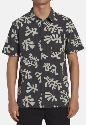 Homme portant une chemise noire à manches courtes avec de grands motifs abstraits de feuilles blanc cassé et une poche poitrine.