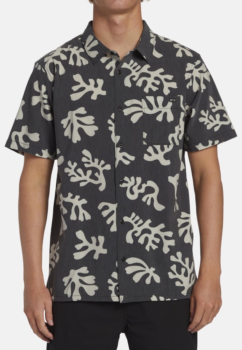 Homme portant une chemise noire à manches courtes avec de grands motifs abstraits de feuilles blanc cassé et une poche poitrine.