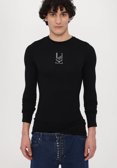 Schwarzes Langarmshirt aus elastischem Stoff, mit einem mit Kristallen besetzten Logodesign auf der Vorderseite, kombiniert mit blauen Jeans.