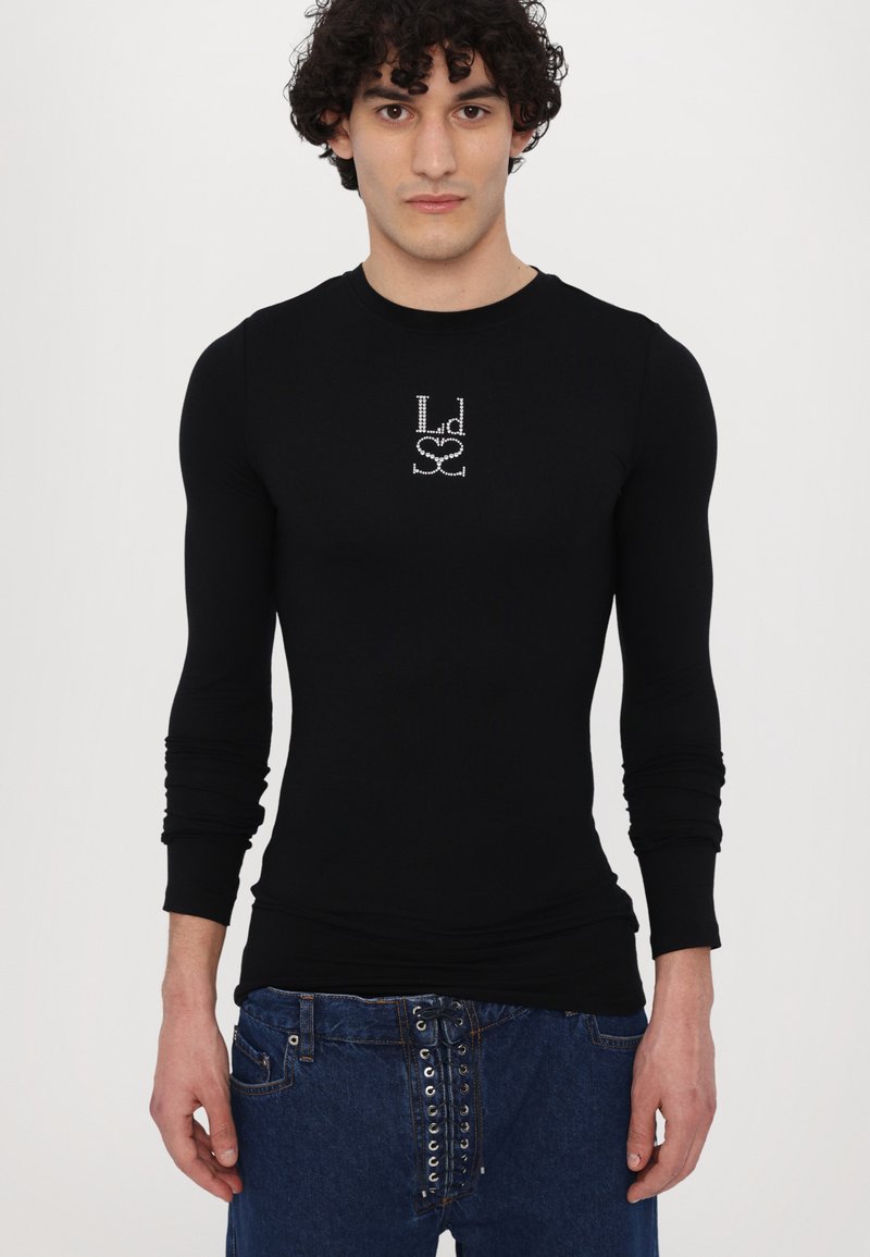 Svart långärmad skjorta i stretchigt material, med en kristallprydd logotypdesign på framsidan, kombinerad med blå denimjeans.