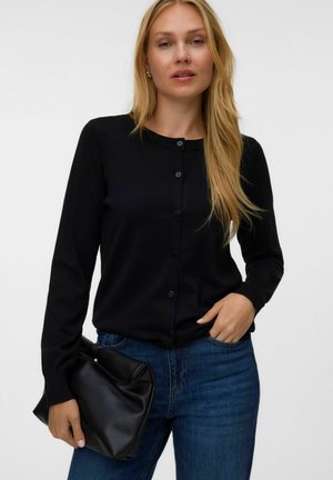 Kvinde med langt blondt hår iført en sort knappet cardigan og blå jeans, der holder en sort foldet læderclutch i den ene hånd.