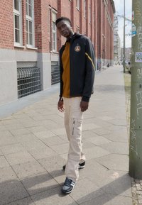 Schwarze Zip-Jacke mit gelben Akzenten, oranges Polo-Shirt, beige Cargo-Hosen mit seitlichen Reißverschlüssen und grau-schwarze Sportschuhe.