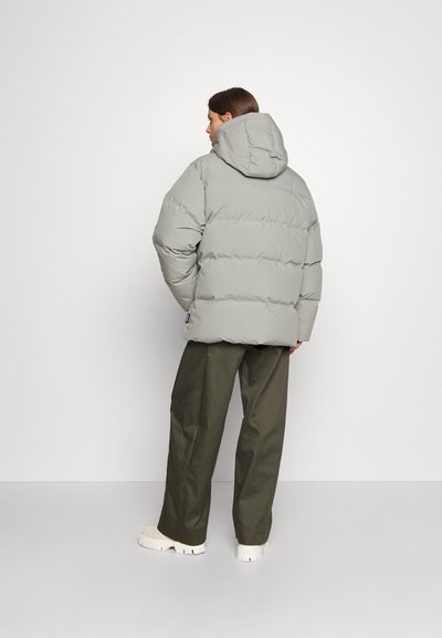 Grå pufferjakke med hætte, med en kviltet design og tyk polstring, parret med olivengrønne, løstsiddende bukser og hvide sko.