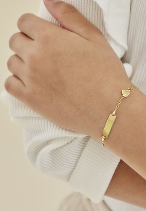 Bracciale d'oro con un ciondolo rettangolare, maglie a catena e un accento decorativo a forma di cuore, indossato su una mano su uno sfondo di tessuto chiaro.