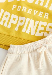 Gelbes Sweatshirt mit weißem Text "Sunshine Forever Happiness" über cremfarbigen gerippten Shorts mit elastischem Bund und Kordelzug.