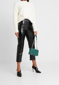Weißer flauschiger Pullover, glänzende schwarze Hose mit Krokodilprägung, grüne strukturierte Handtasche mit Kettenriemen und schwarze spitze Stilettos.