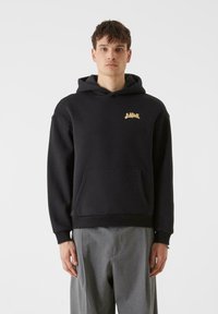 Črna kapuca iz mehkega blaga, z sprednjim žepom in zlatim logotipom na prsih. Oblikovana z udobnim krojem in z zategovanjem na kapuci.